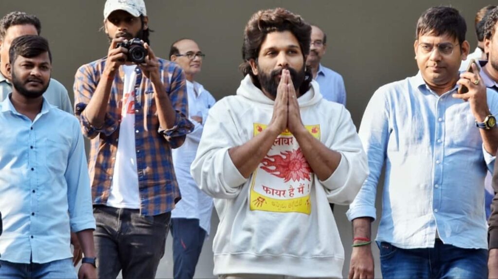 A man with folded hands and a white hoodie at a public event, surrounded by supporters and photograp.