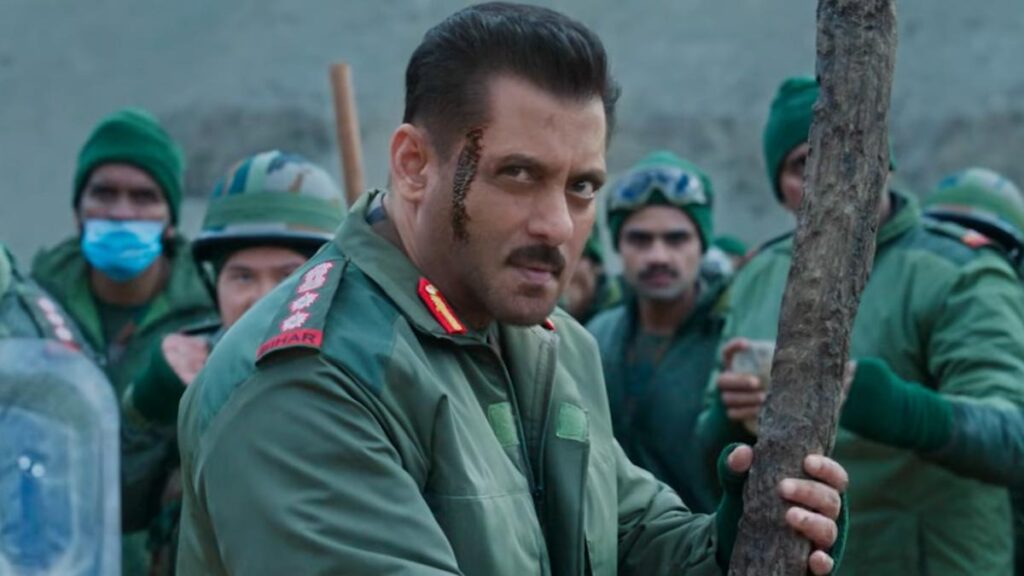 Indian military officer with soldiers in the background, outdoor setting, serious expression.