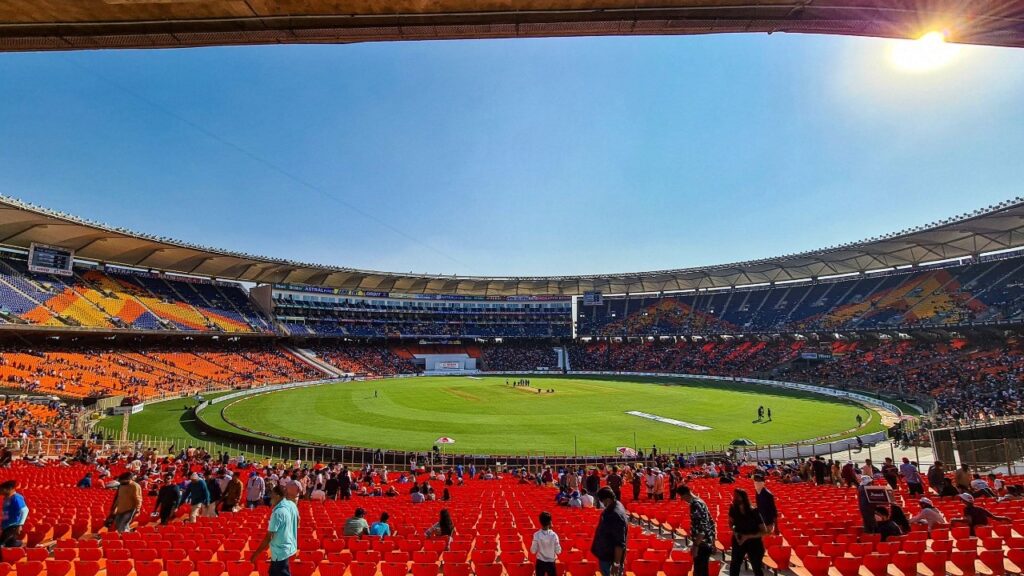 View of a large outdoor stadium with a cricket field and seating area during daytime.