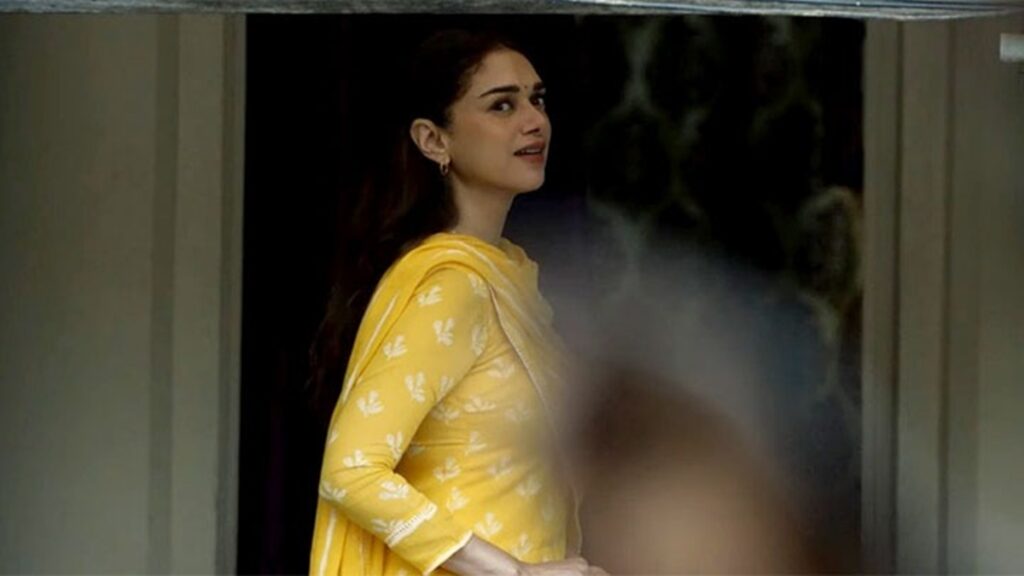 A woman in a yellow traditional outfit looking out of a window with steam or smoke in the background.