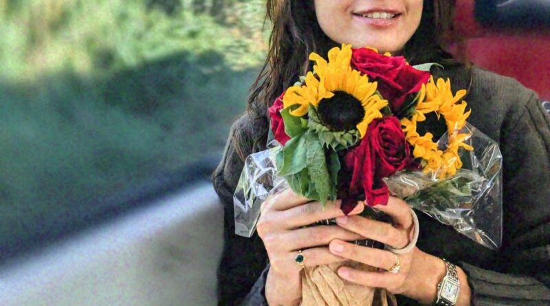 Sunflower and rose bouquet held by a smiling woman with glasses on a bus.